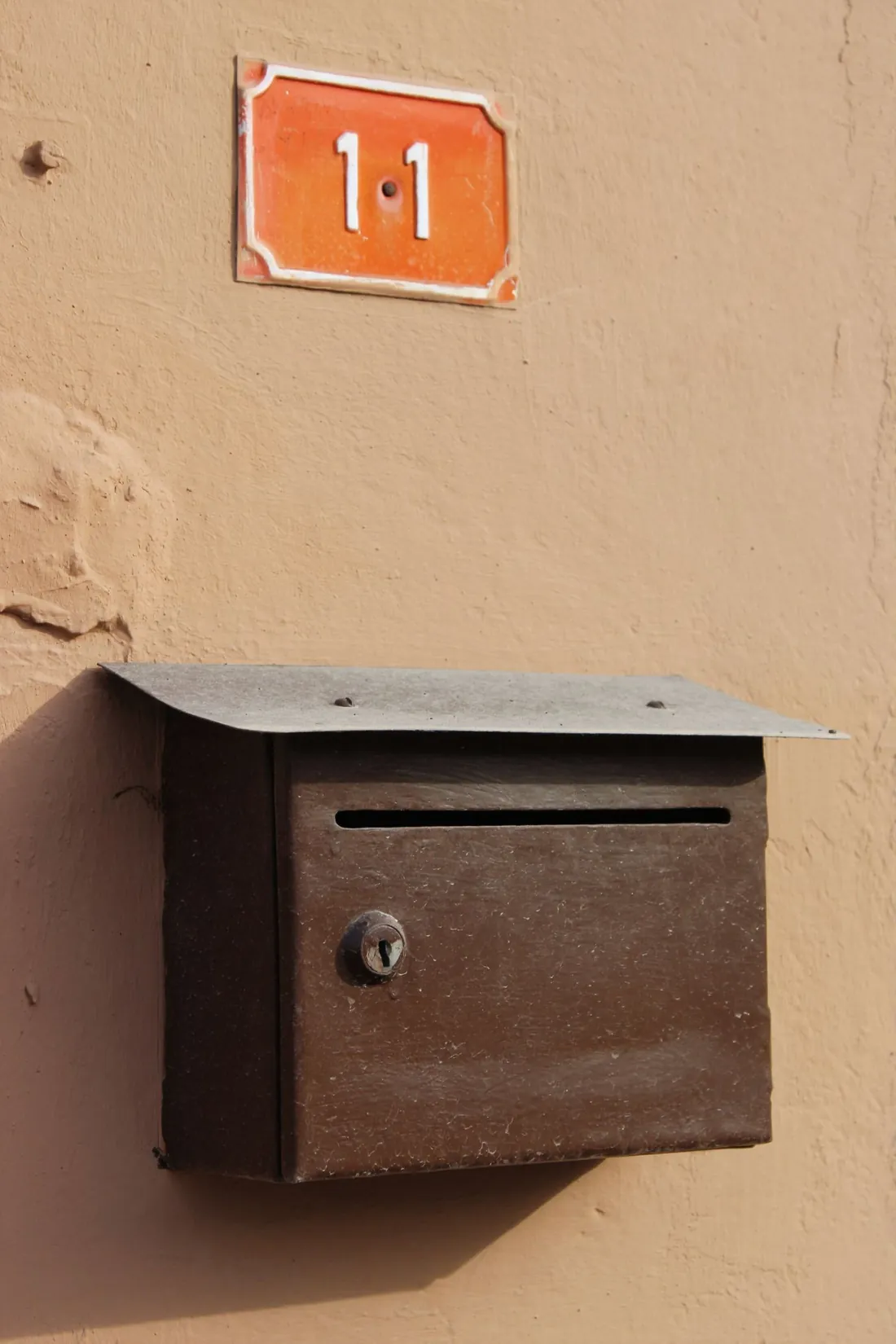 Boîte aux lettres métallique fixée sur un mur ocre, sous une plaque de numéro de rue émaillée