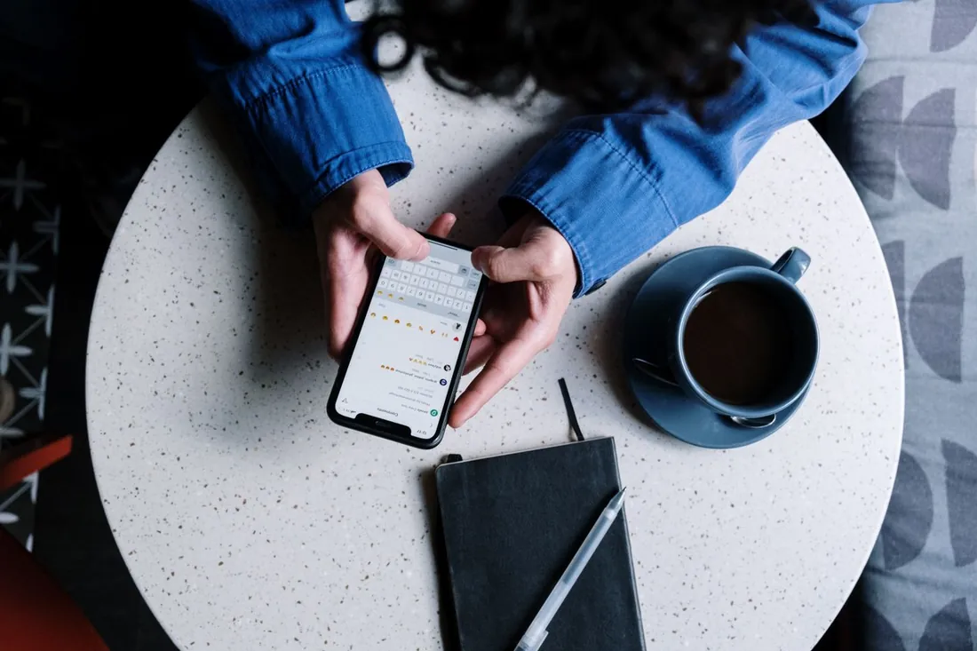 Vue de dessus d'une table de café : deux mains tiennent un téléphone, un carnet et une tasse posés à côté
