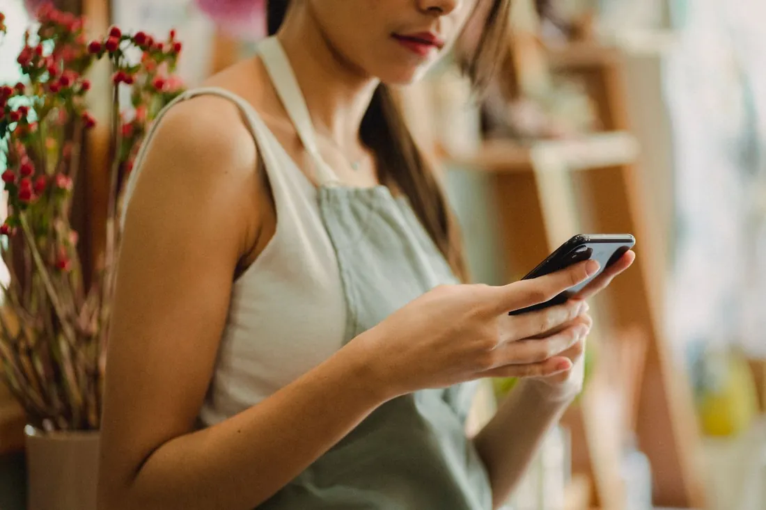 Fleuriste en tablier dans sa boutique, qui regarde son site sur son téléphone entre deux clients
