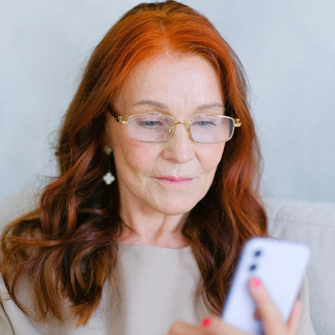 Femme aux cheveux roux et aux lunettes qui lit l'écran de son téléphone en baissant les yeux