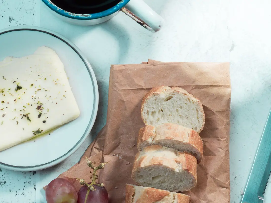 Petit-déjeuner rustique vu de dessus : café noir, baguette tranchée, fromage et raisin sur une table claire