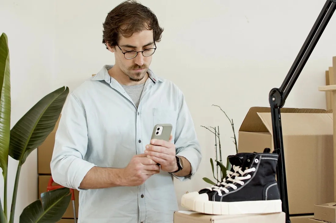 Artisan qui photographie un de ses produits avec son téléphone dans son atelier, cartons et lampe autour de lui