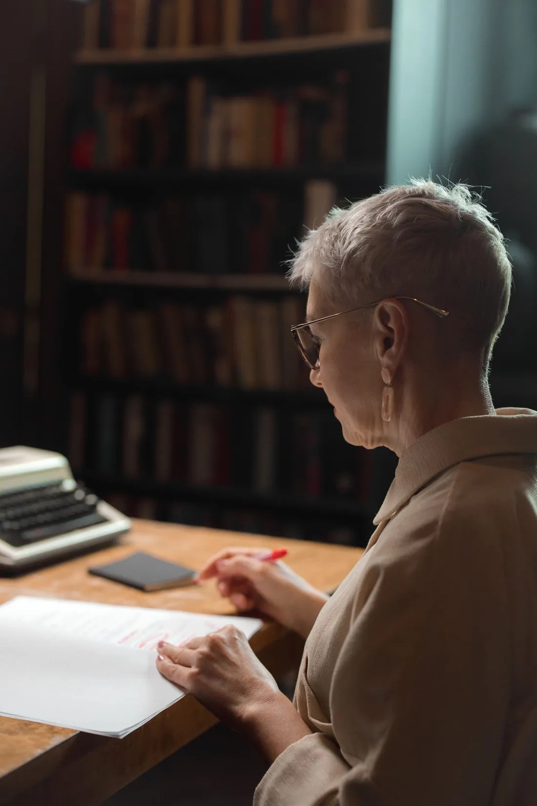 Woman with short hair sitting at a library desk, red pen in hand, re-reading a printed text