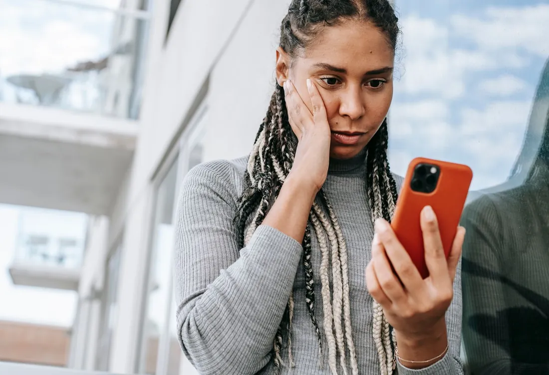 Femme dans la rue, la main sur la joue, qui hésite devant l'écran de son téléphone après une recommandation